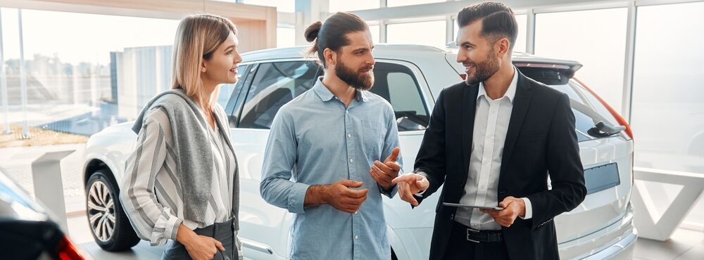 Car salesman in a suit talks with a smiling couple inside a dealership, gesturing while holding a tablet. A white SUV is displayed behind them