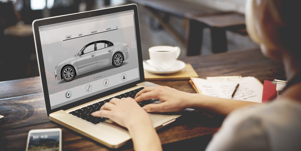 Person browses a car shopping website on a laptop, viewing a silver sedan. A cup of coffee, a notepad, and a smartphone are on the wooden table