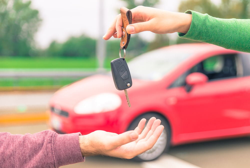 Person in a green sleeve hands over a car key to another person's open palm, with a red car parked in the background