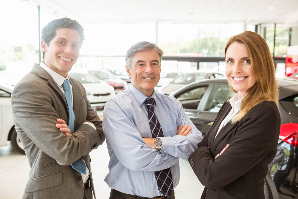 Three smiling car salespeople stand with arms crossed inside a dealership, surrounded by various cars in a bright showroom